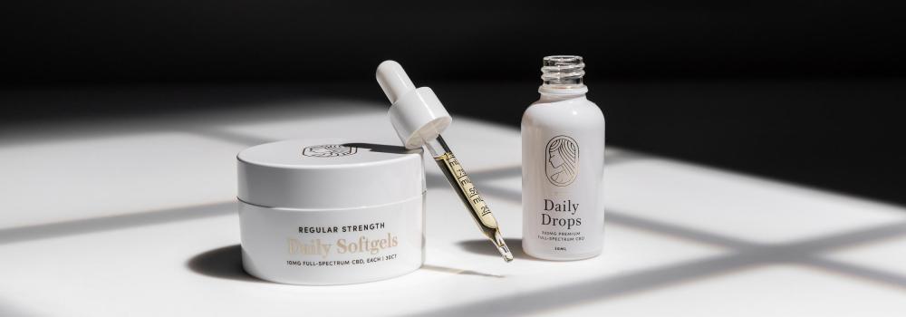 Regular Strength Dail Soft Gels next to open bottle of regular strength Daily Drops. Full dropper of Daily Drops leaning on Daily Softgels container. White surface with dramatic shadows.