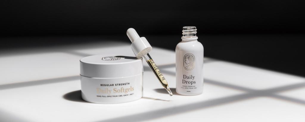 Regular Strength Dail Soft Gels next to open bottle of regular strength Daily Drops. Full dropper of Daily Drops leaning on Daily Softgels container. White surface with dramatic shadows.